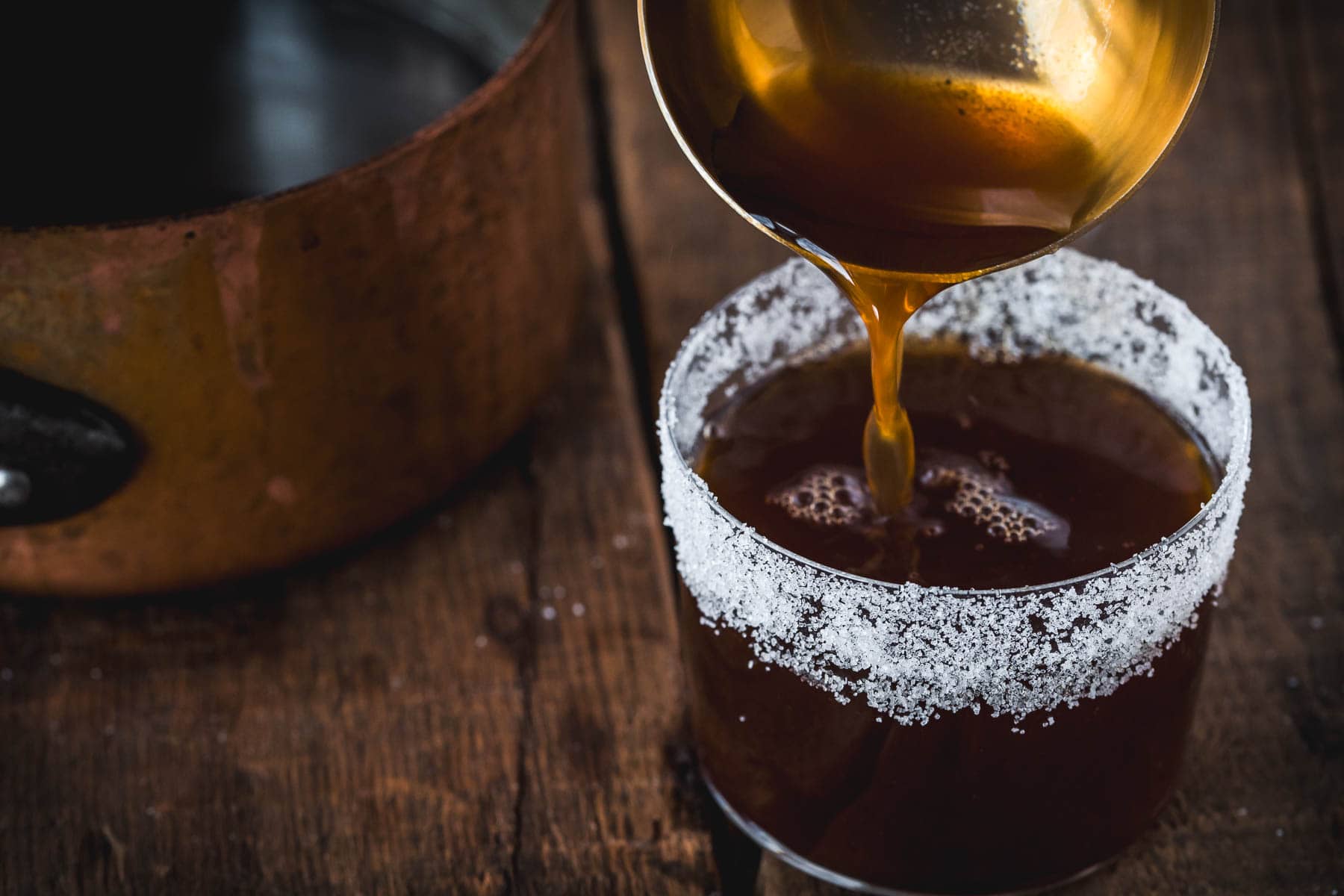 A golden ladle pours a dark liquid into a sugar-rimmed glass, crafting a Colombian Canelazo cocktail. The glass rests on a wooden surface, with a copper pot partially visible in the background.