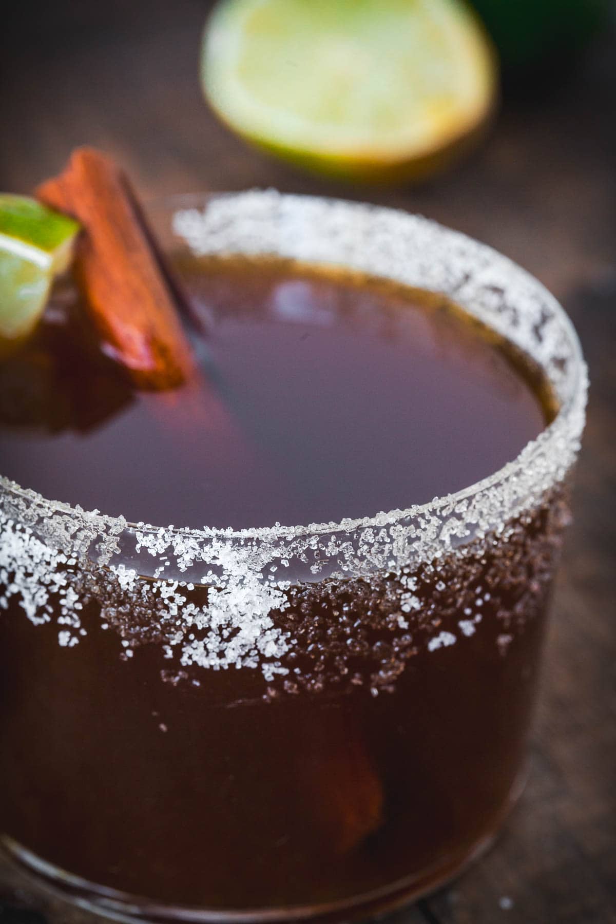 A close-up of a glass with a salted rim filled with a dark brown Colombian Canelazo cocktail, garnished with a lime wedge and cinnamon stick. A lime half is blurred in the background on a wooden surface.