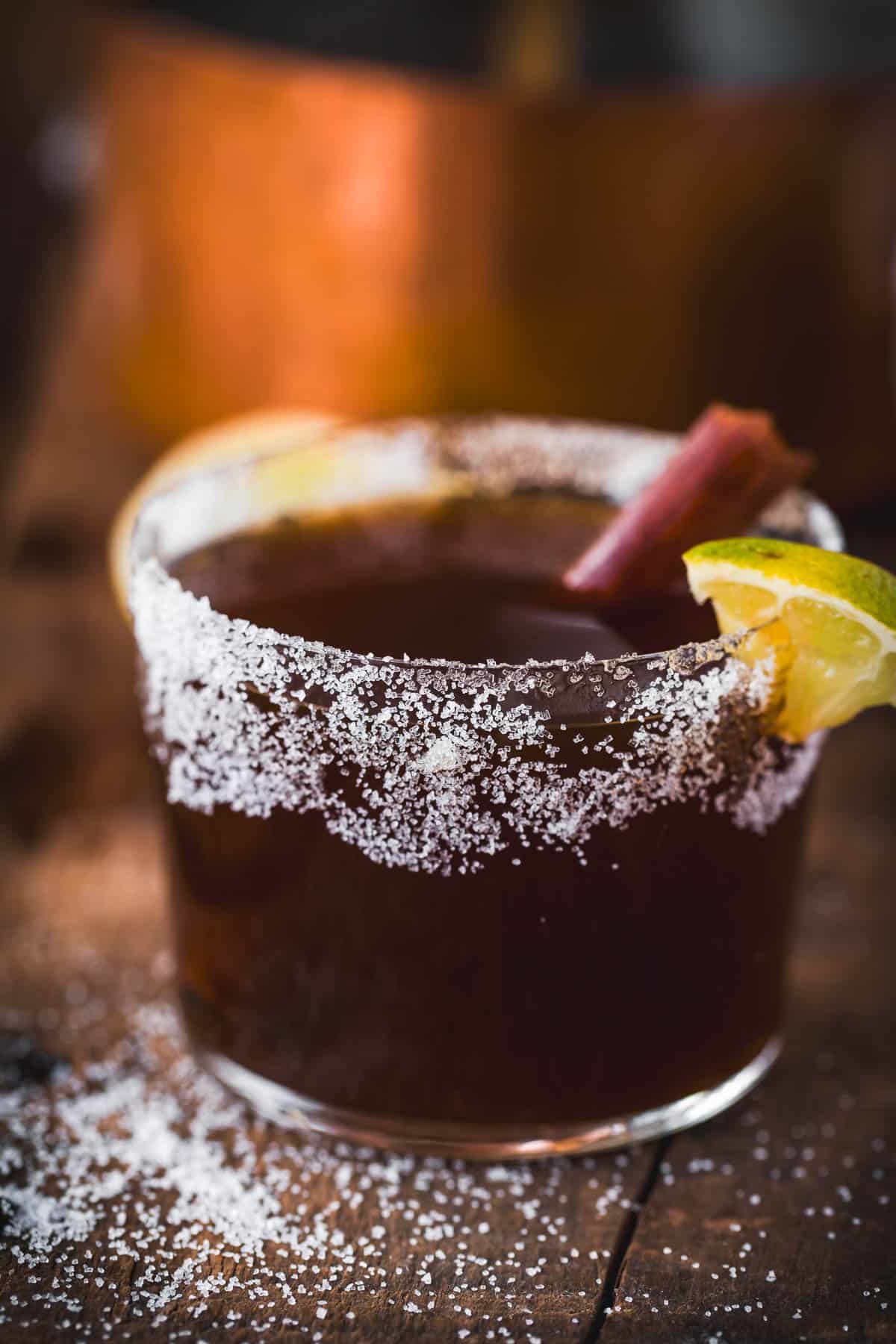 A glass filled with a dark Colombian Canelazo, rimmed with coarse salt and garnished with a lime wedge and stick, sits on a wooden surface sprinkled with salt crystals.