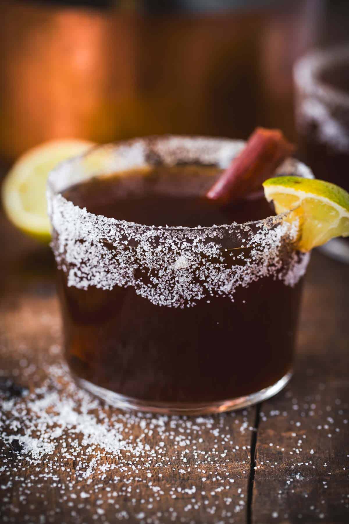 A brown Canelazo Cocktail in a glass with a salted rim, garnished with a lemon wedge, lime wedge, and cinnamon stick, sits on a wooden surface sprinkled with salt. Another Colombian cocktail is blurred in the background.