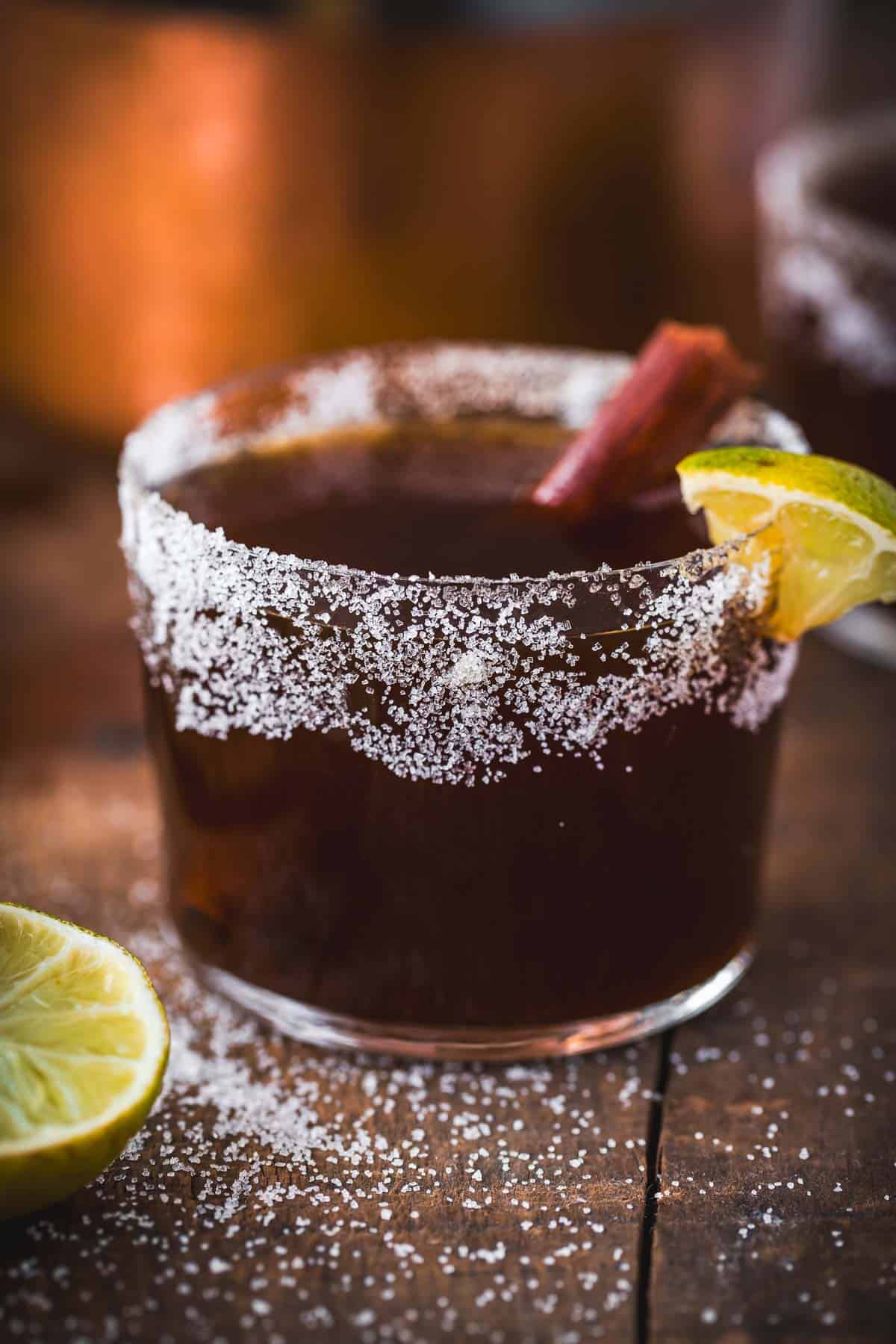 A glass filled with a dark Colombian Canelazo cocktail, rimmed with salt and garnished with a lime wedge and a stick, sits on a wooden surface with sliced lime and scattered salt nearby.
