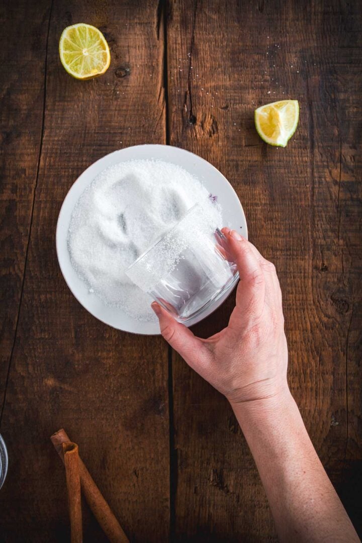 A hand holds a glass, pressing its rim into a plate of sugar on a wooden surface—prepping for a Colombian Canelazo cocktail. Two lemon wedges rest on the table nearby.