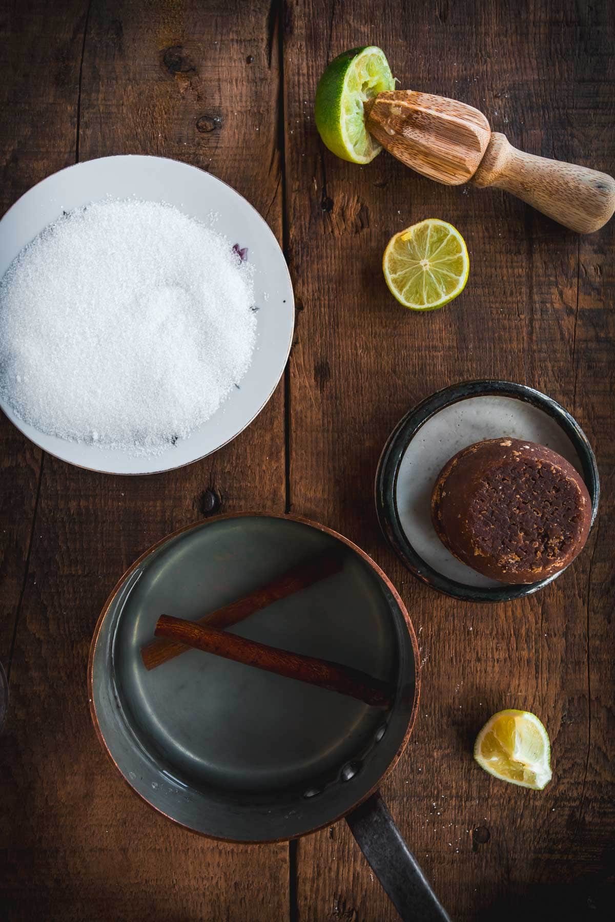 A saucepan with water and two cinnamon sticks sits on a wooden surface next to a bowl of sugar, a round block of brown sugar, a citrus reamer, and cut limes—perfect ingredients for making a traditional Colombian Canelazo cocktail.
