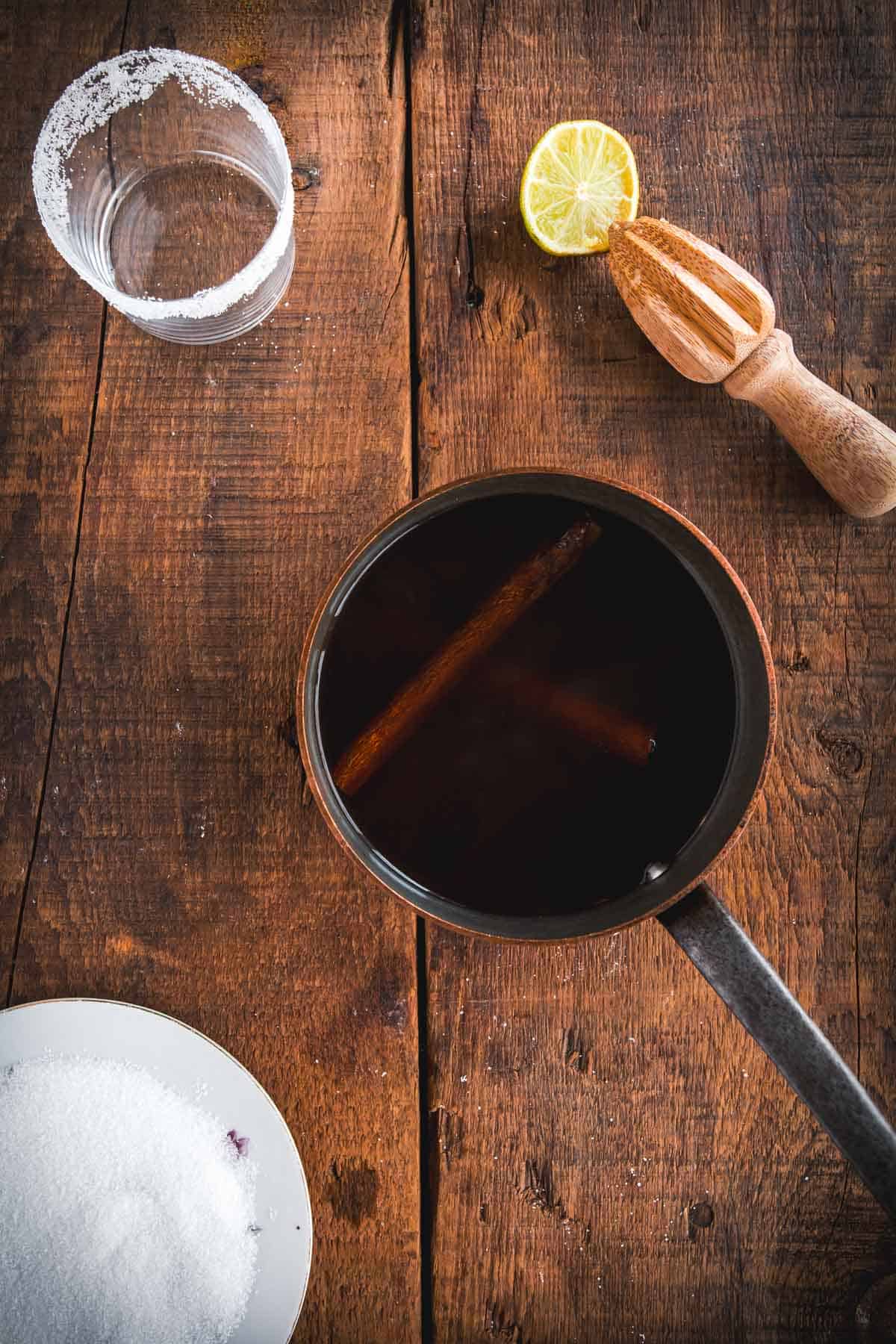A saucepan with dark liquid and cinnamon sticks sits on a wooden surface. Nearby are a halved lemon, a wooden citrus juicer, a salt-rimmed glass, and a plate of white granulated sugar—ingredients for an authentic Colombian Canelazo cocktail.