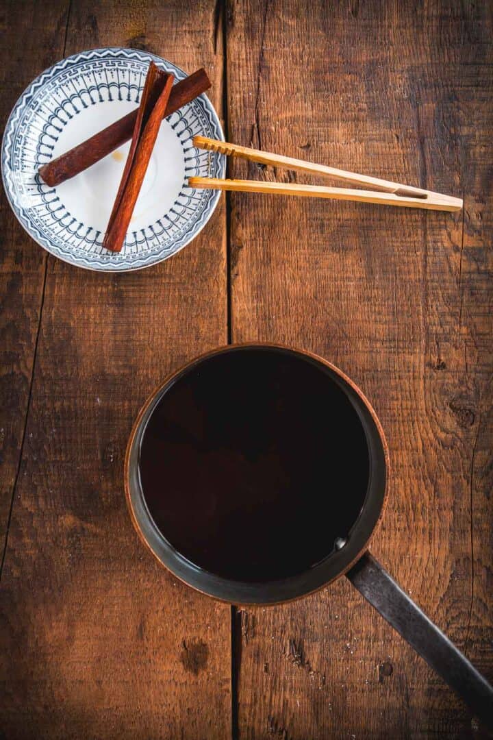 A white patterned bowl with cinnamon sticks and wooden tongs sits on a wooden surface next to a pot filled with dark Colombian Canelazo, viewed from above.