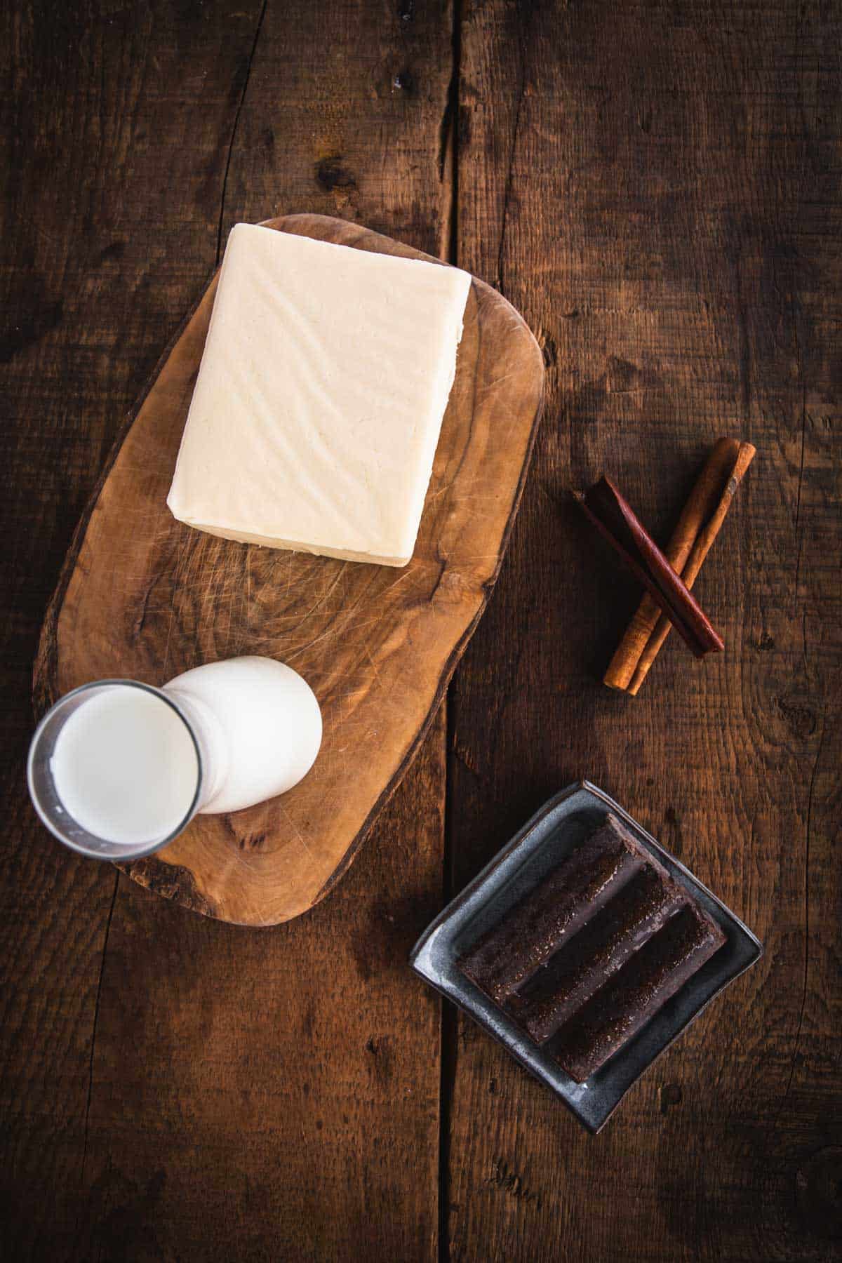 A block of white cheese on a wooden board, a glass of milk, cinnamon sticks, and a small tray of brown sticks sit on a rustic wooden surface, reminiscent of cozy mornings with Colombian hot chocolate.