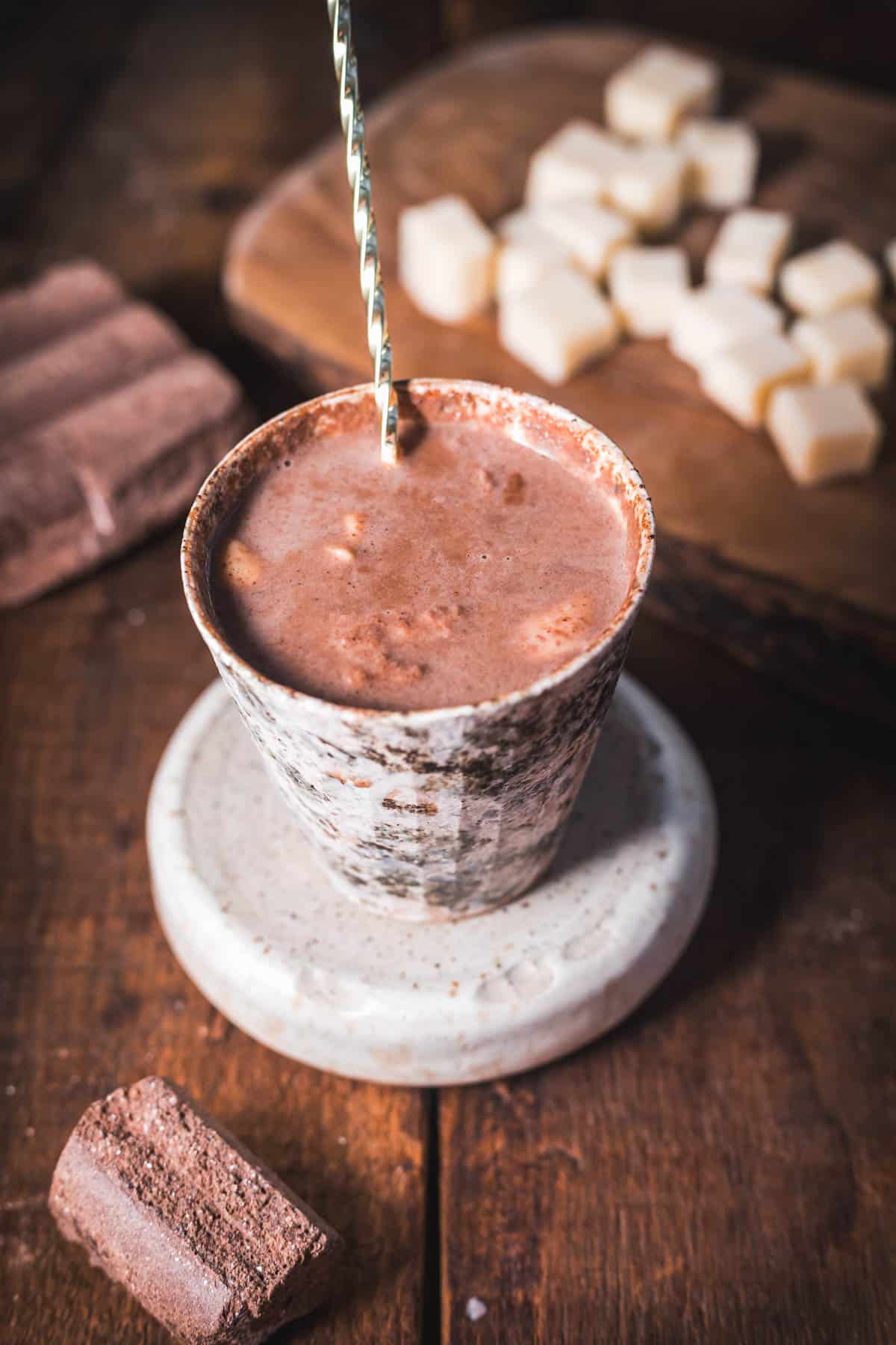 A ceramic cup filled with rich Colombian hot chocolate rests on a round coaster, a silver spoon inside. In the background, cube-shaped pieces of cheese and chocolate bars sit enticingly on a rustic wooden surface.