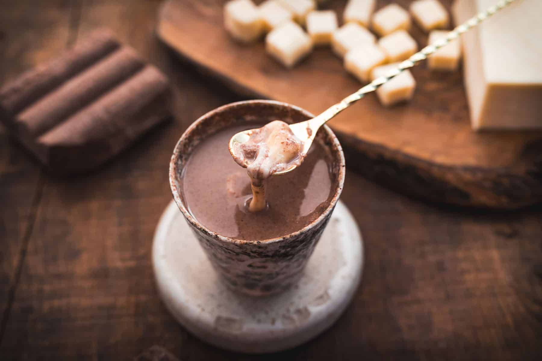 A rustic ceramic cup filled with Colombian hot chocolate on a saucer, with a spoon lifting a dollop from the cup. In the background, there are wooden blocks and small cubes on a wooden board, suggesting additional chocolate pieces, enhancing the rich experience.