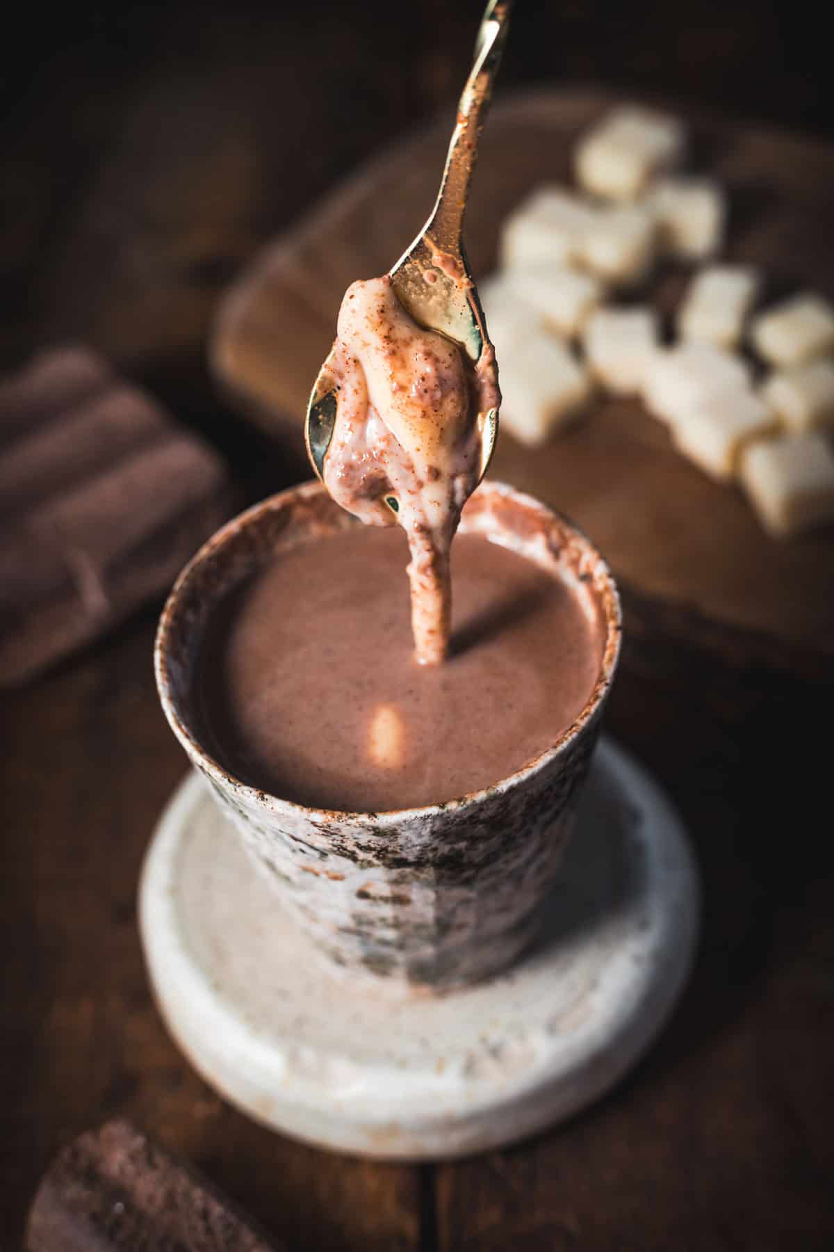 A ceramic cup filled with Colombian hot chocolate sits on a saucer. A spoon dips in, lifting a dollop of melting marshmallow. In the background, a wooden board holds sugar cubes and a chocolate bar, completing the quintessential hot chocolate recipe scene.