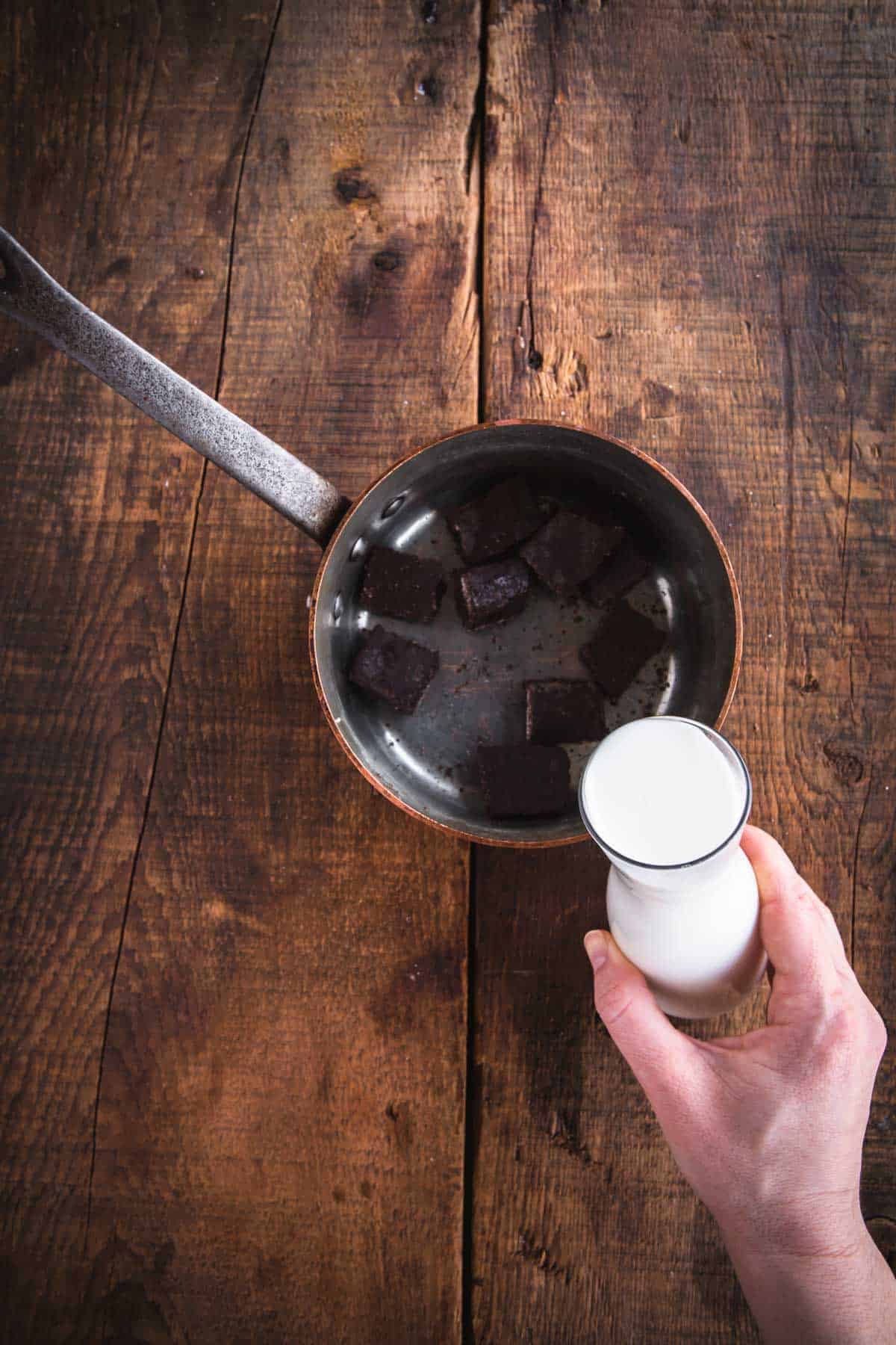 A hand holds a small glass bottle of milk above a saucepan with dark Colombian chocolate pieces, ready to craft a rich hot chocolate on the rustic wooden surface.