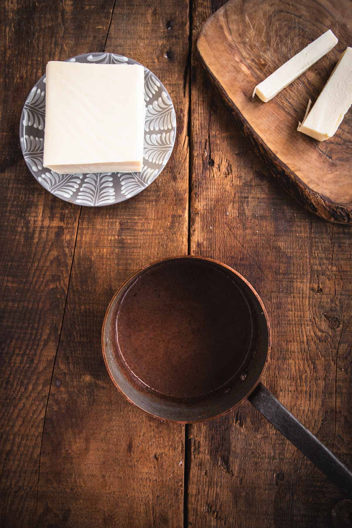 A rustic wooden table displays an empty brown pan with a long handle, perfect for preparing Colombian hot chocolate. To the top left, a decorative plate holds stacked white cheese slices. To the top right, a wooden board showcases cut cheese pieces, ready to be paired with the rich beverage.