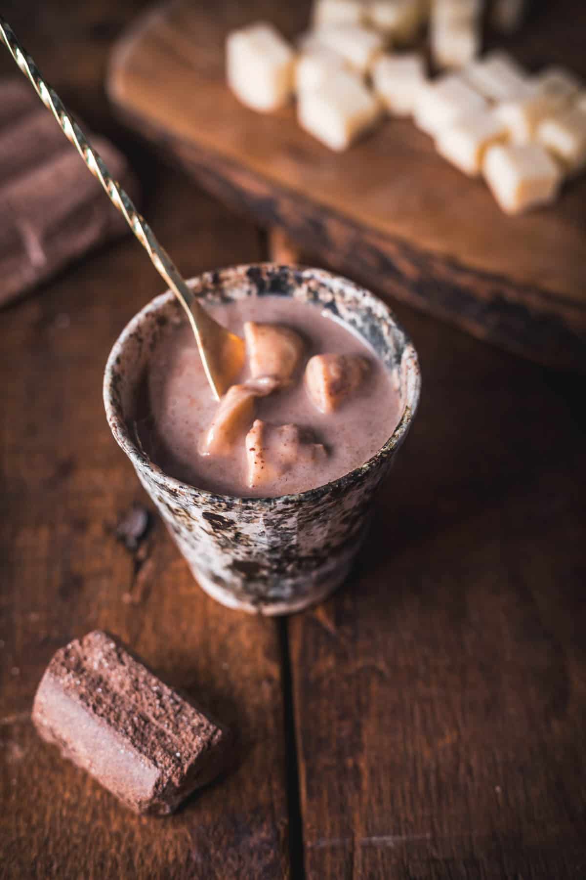 A cup of Colombian hot chocolate with marshmallows sits on a wooden table, complemented by a gold spoon glistening in the rich brew. In the background, cubes of cheese rest on a wooden board alongside a piece of dark chocolate.