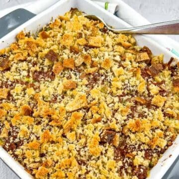 A square baking dish filled with a golden-brown casserole topped with crumbled bread. A serving spoon rests inside the dish. The dish is placed on a white tray, with a floral-patterned napkin nearby.