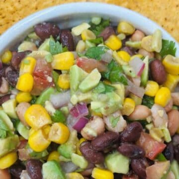 A bowl of bean salad containing black beans, corn, avocado, diced tomatoes, onions, and cilantro, surrounded by tortilla chips.