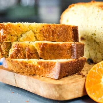Three slices of pound cake are stacked on a wooden cutting board next to the remaining loaf. Two halved oranges are placed beside the cake, and a blue-and-white cloth is partially visible in the background.