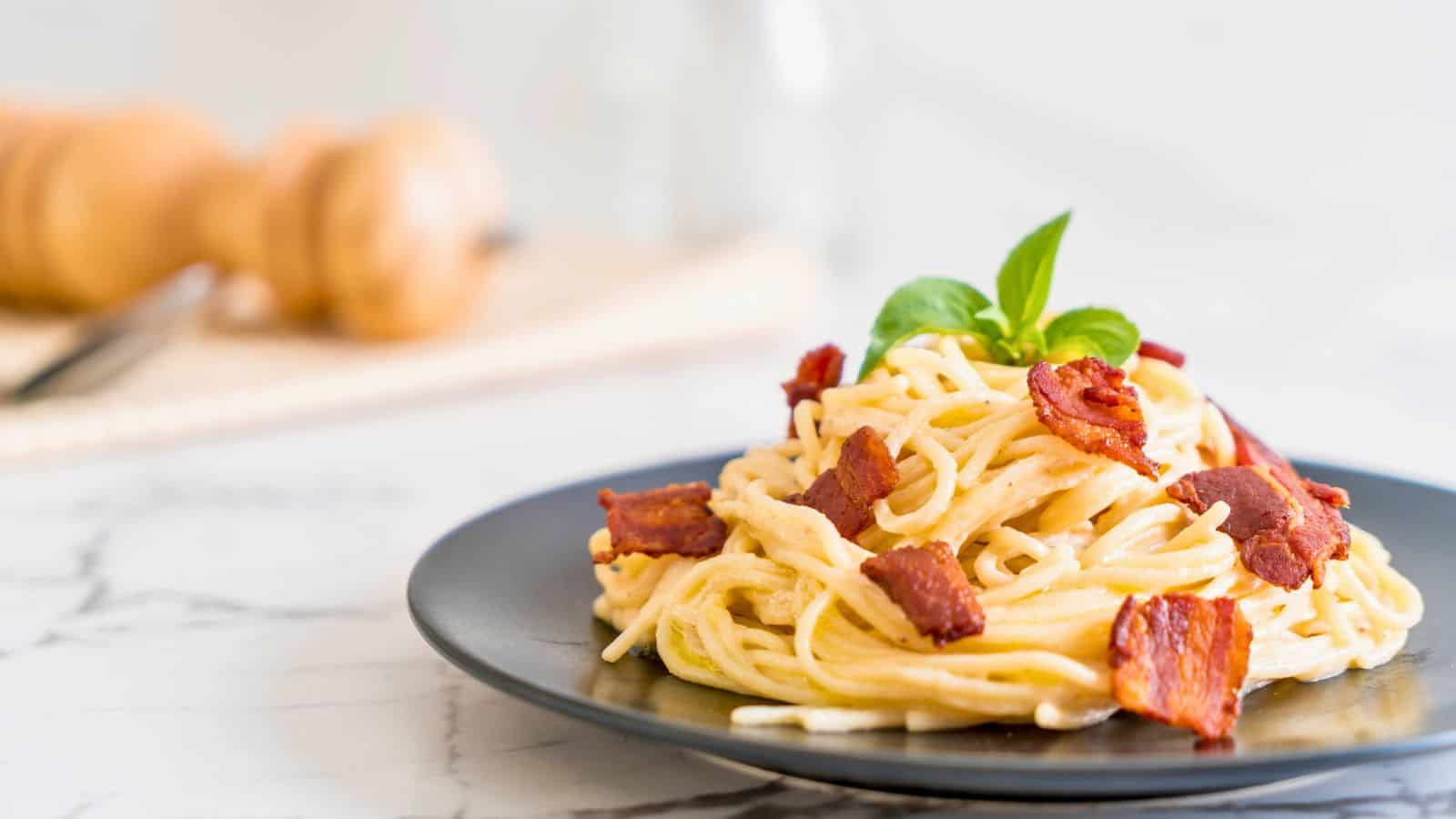 A plate of creamy spaghetti carbonara topped with pieces of crispy bacon and a basil garnish. The dish is served on a black plate, set against a marble surface, with a pepper grinder blurred in the background.