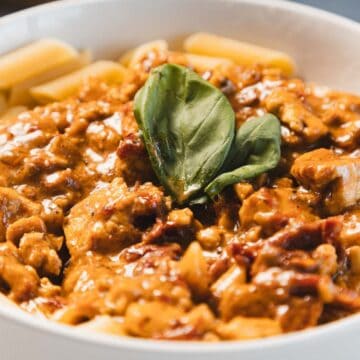 A bowl of creamy sun-dried tomato tofu sauce blankets chunks of meat, with fresh basil leaves as a garnish. The rigatoni, nestled perfectly in the rich Marry Me Tofu blend, is served elegantly in a pristine white bowl.