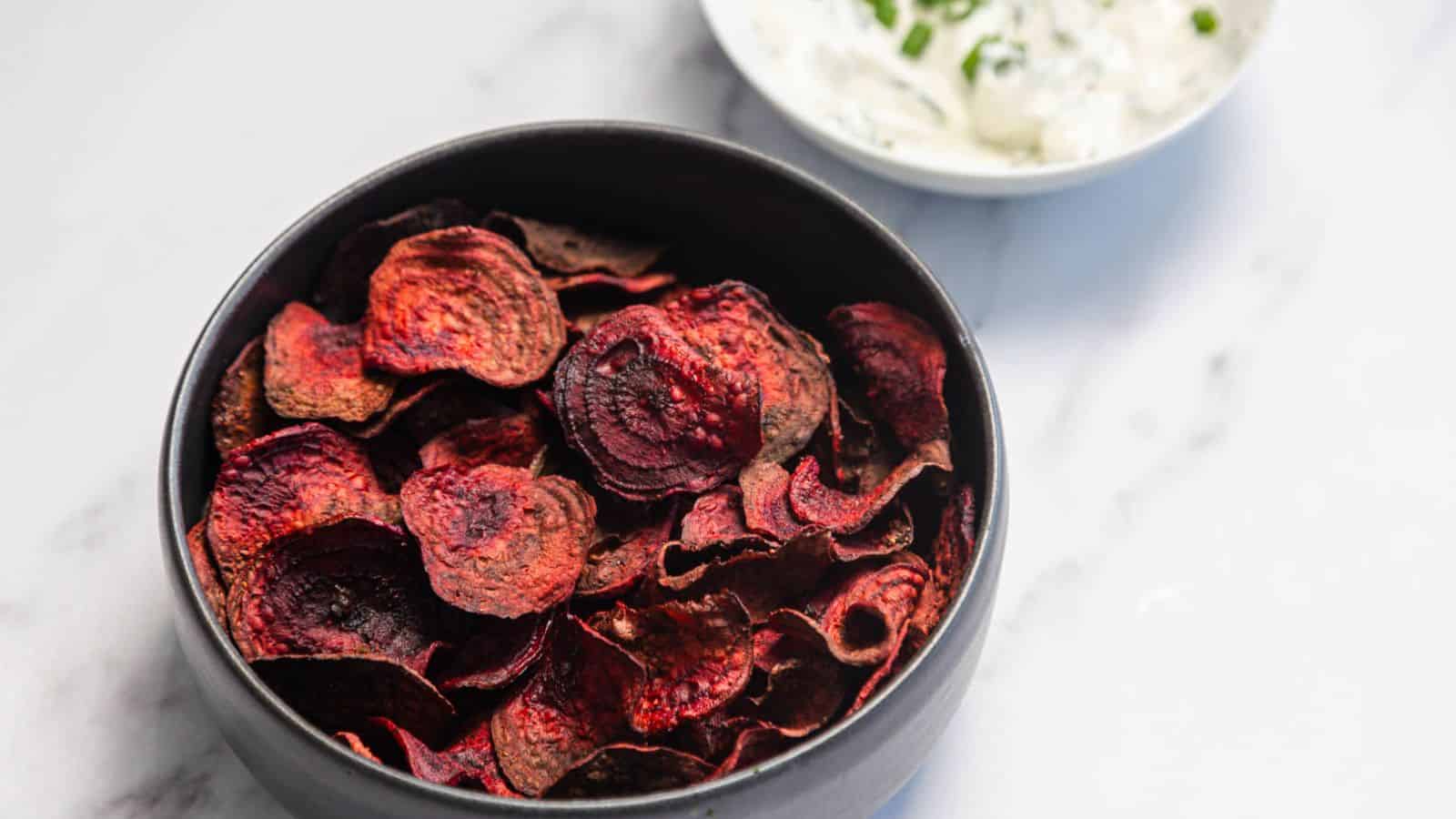 A black bowl filled with dark red beet chips sits on a white surface. In the background, there is a small white bowl containing a creamy dip with green herbs.