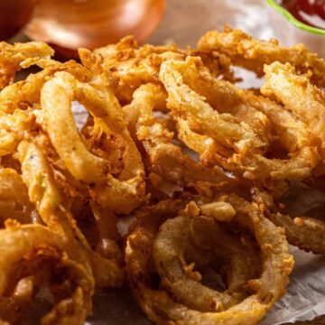 A close-up of crispy fried onion rings on parchment paper, with a bowl of red ketchup and whole onions in the background.