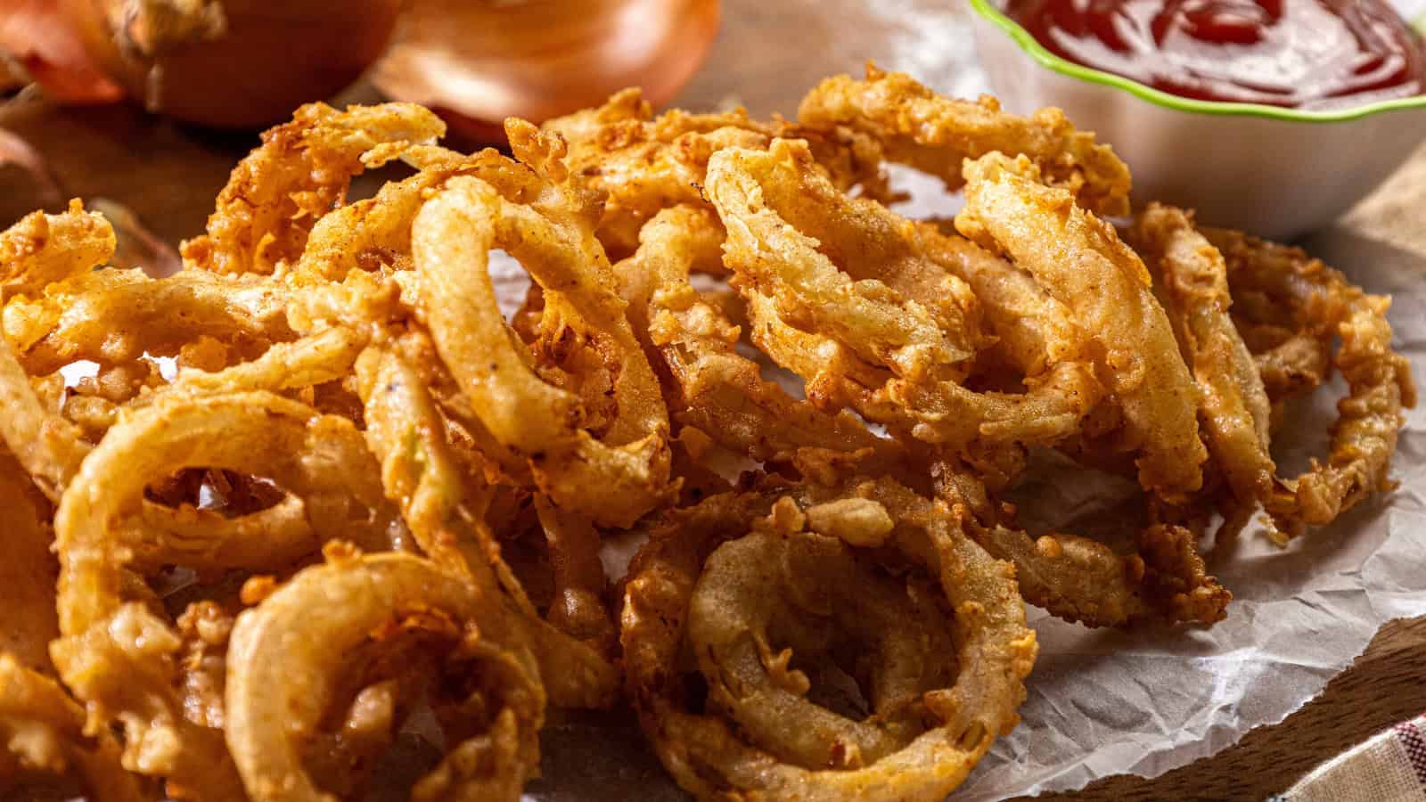 A pile of crispy fried onion rings rests on parchment paper, accompanied by a small bowl of red dipping sauce in the background.