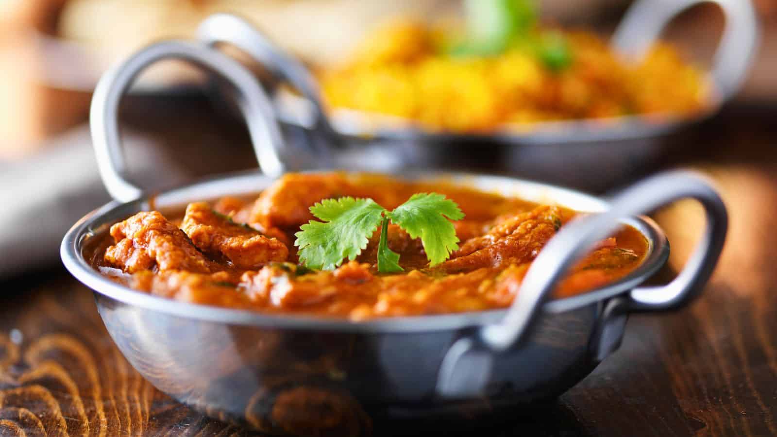 A stainless steel bowl filled with a spicy curry, garnished with cilantro leaves. The curry contains pieces of meat or vegetables in a rich, reddish-brown sauce. Another dish, possibly rice, is blurred in the background.