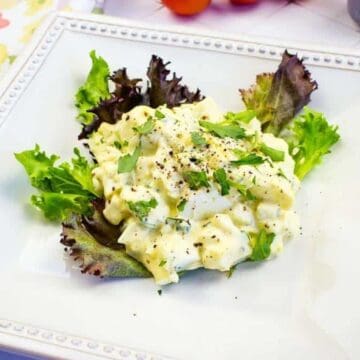 A square white plate with egg salad, reminiscent of retro recipes, garnished with parsley and black pepper, rests on a bed of mixed greens. Part of a floral tablecloth adds charm in the corner—perfect for an Instagram story comeback.