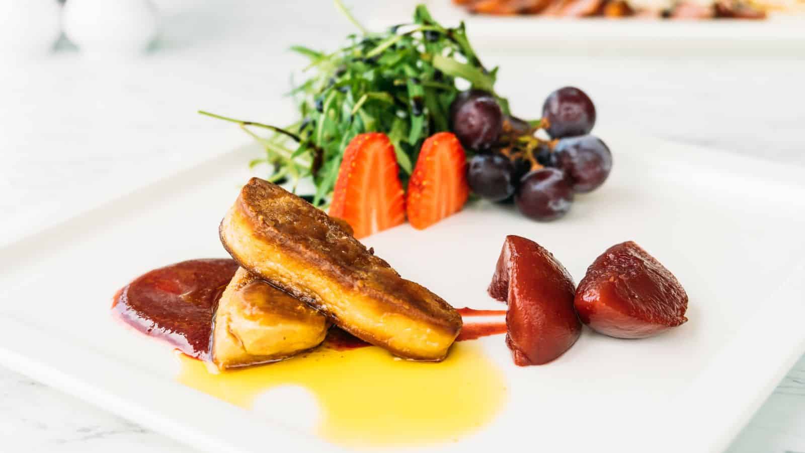 A gourmet dish on a white plate features two pieces of foie gras with sauce, accompanied by poached pear slices, fresh strawberries, red grapes, and a small arugula salad.
