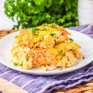 A white plate holds a serving of cheesy baked casserole garnished with chopped herbs. The plate sits on a blue-striped cloth next to a wooden spoon, with an onion, green parsley, and a drink in the background.