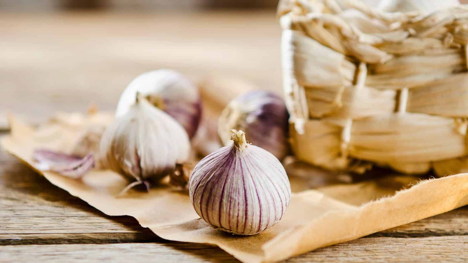 Three bulbs of garlic with purple streaks on a piece of brown parchment paper, beside a woven basket. The scene is set on a wooden surface.