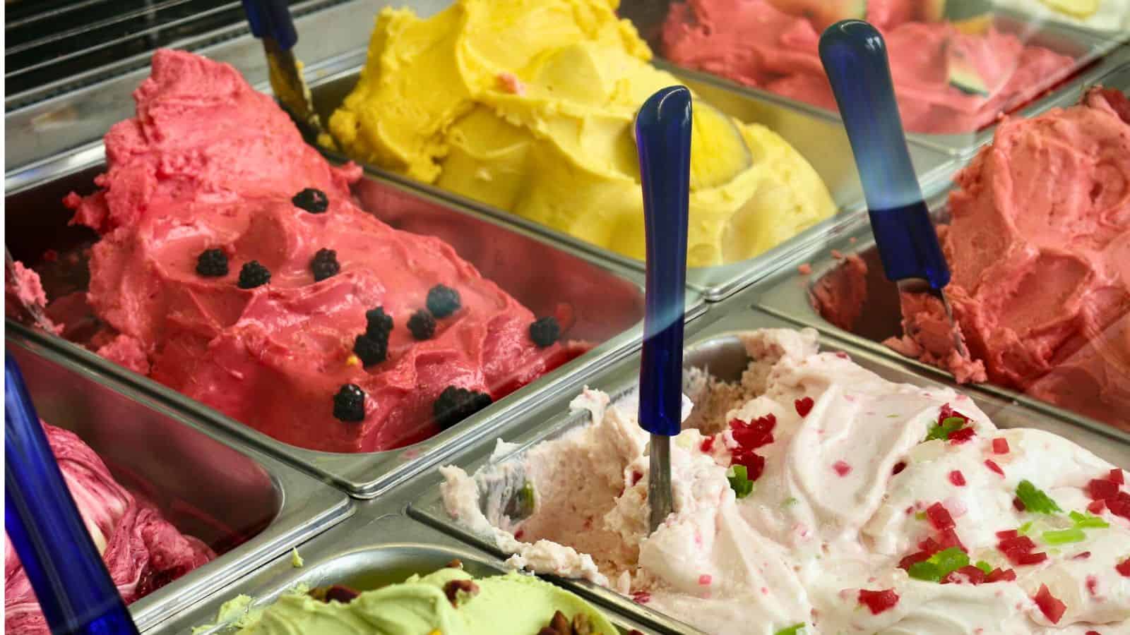 A display of various gelato flavors in metal containers, each with a blue-handled scoop. Visible flavors include pink with berries, yellow, red, pale pink with fruit toppings, and light green with nuts.