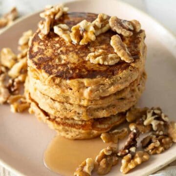 A stack of four pancakes topped and surrounded by walnuts and drizzled with syrup sits on a light pink plate. A cup of coffee and a fork are slightly visible in the background.