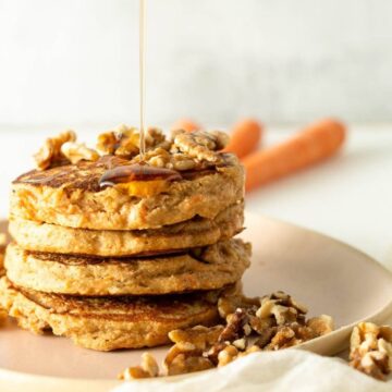 A stack of three pancakes topped with walnuts is on a pink plate. Syrup is being drizzled over the pancakes. Additional walnuts are scattered around the plate. Two carrots are partially visible in the background.
