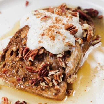 A slice of French toast topped with whipped cream, chopped pecans, and a sprinkle of cinnamon, served on a white plate with syrup pooling around it.