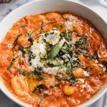 A white bowl filled with tomato-based gnocchi topped with creamy cheese, chopped fresh basil, and a drizzle of olive oil sits on a white surface with a fork, napkin, and basil leaves nearby.