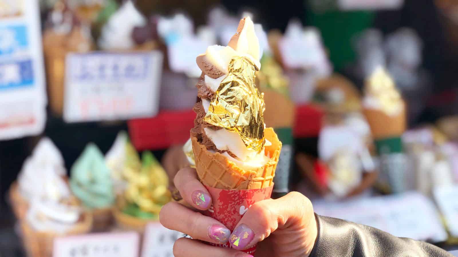 A hand holding a fancy ice cream cone with multiple swirled flavors and gold leaf on top. The cone is decorated with a red sleeve, and the person's nails are intricately painted. The background shows an out-of-focus display of more treats.