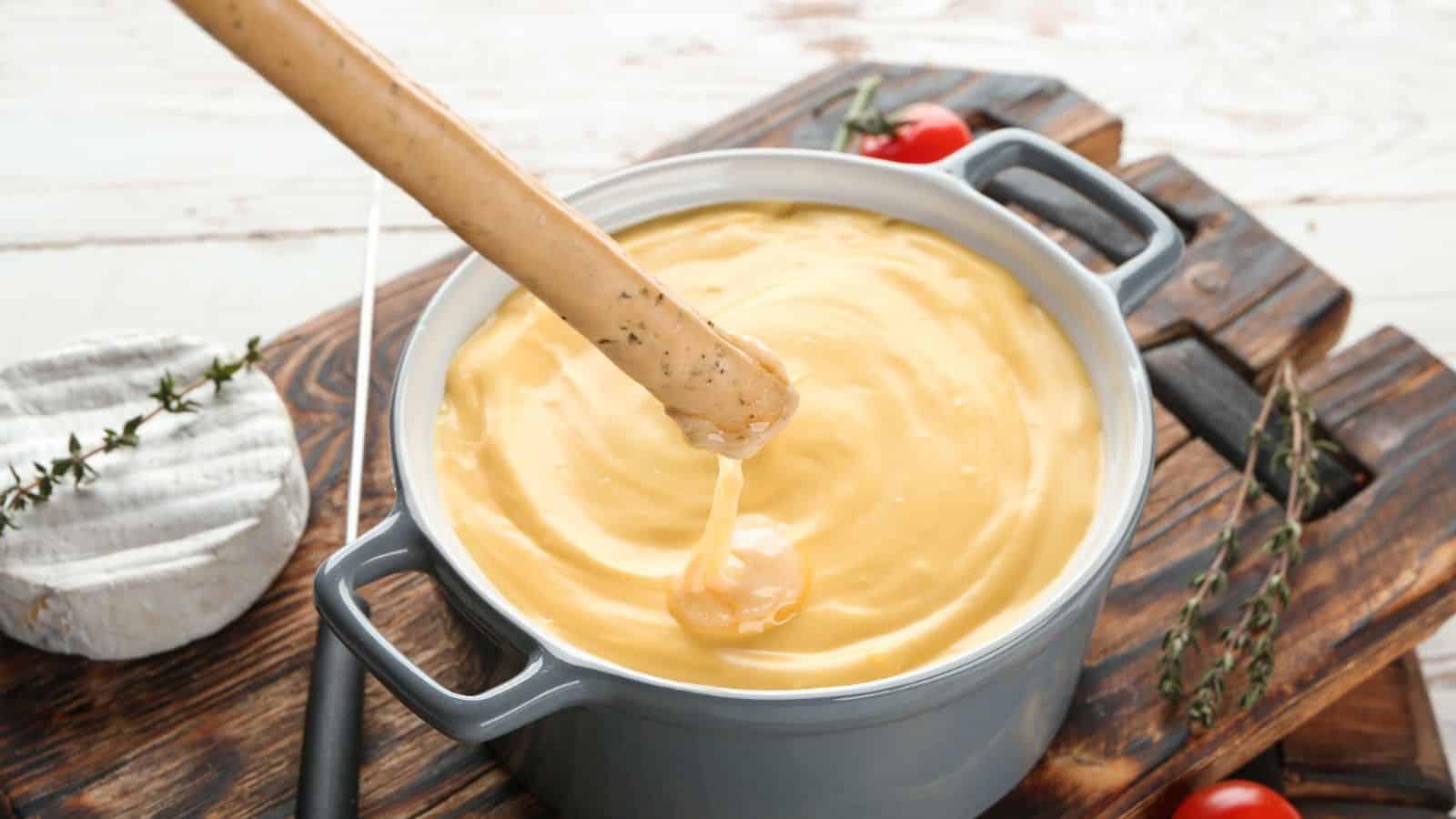 A piece of breadstick is being dipped into a pot of creamy cheese fondue. The fondue pot is on a wooden board next to some cherry tomatoes and sprigs of thyme. A wheel of soft cheese is partially visible on the board.