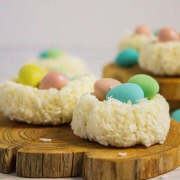 Wooden boards display coconut nests filled with colorful candy eggs. The nests are white, textured with shredded coconut, and contain pastel-colored eggs in shades of pink, blue, and yellow. The background is softly blurred.