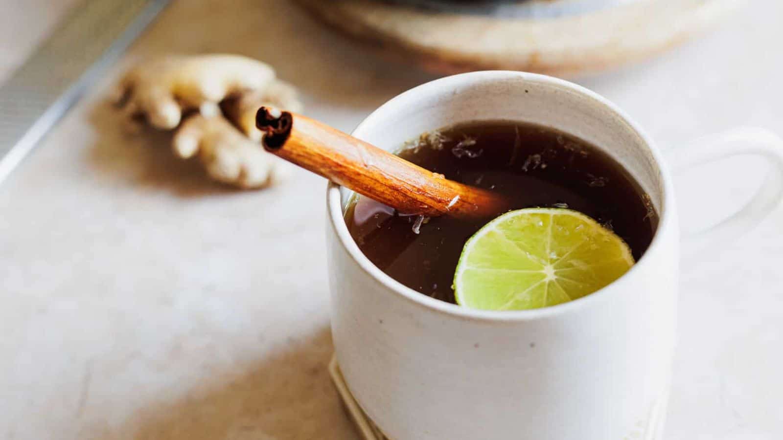 A mug filled with a dark beverage, garnished with a lime slice and a cinnamon stick, sits on a table. Next to it, a small plate holds several round cookies. A piece of ginger is visible in the background.