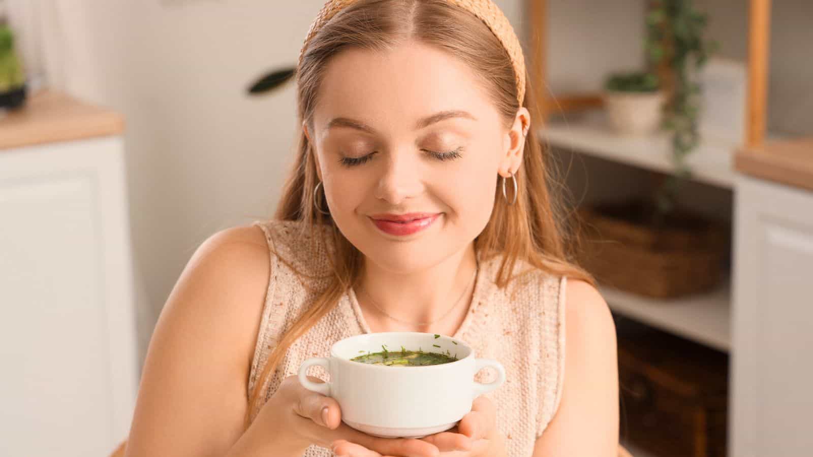 A person with long hair and a headband is seated indoors, holding a bowl of green soup with both hands. They have a content expression, eyes closed, and appear to be savoring the aroma. The background includes shelves and plants.