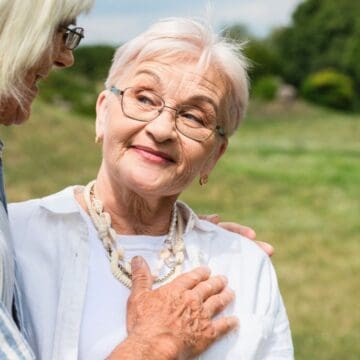 Two elderly women are outdoors in a green field. One faces the camera, smiling, with short white hair and glasses, wearing a white shirt and necklace. The other, with long white hair and a striped shirt, faces her, hand resting gently on her shoulder.