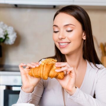 A person with long hair is sitting at a kitchen table, smiling while holding a croissant. Behind them is a vase with flowers, kitchen utensils, and a stove. A glass pitcher and cup are on the table.