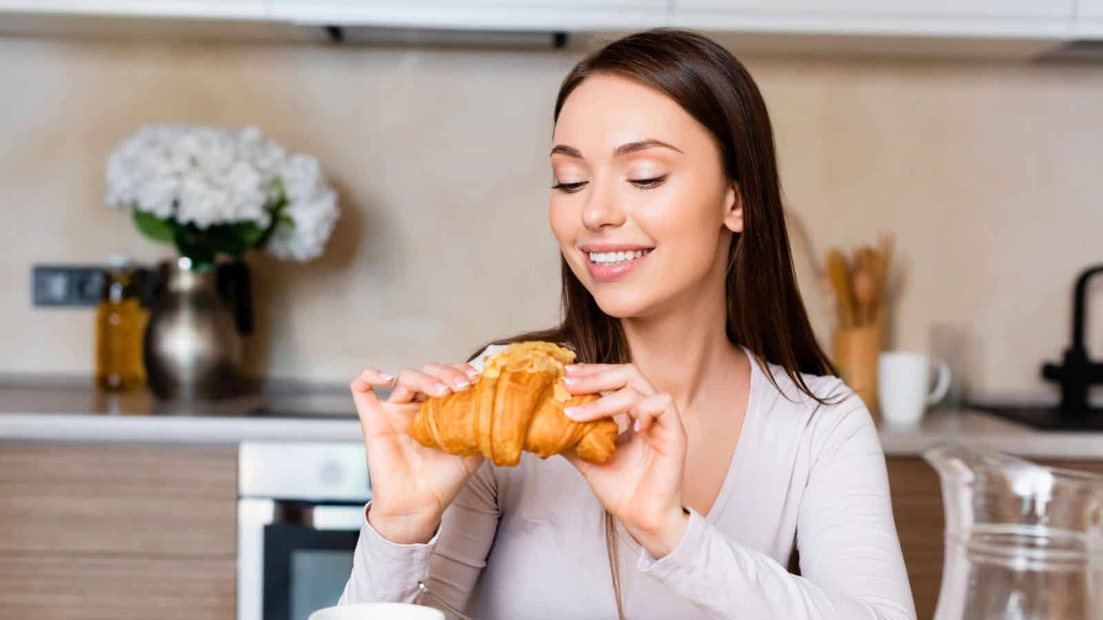 A person with long hair is sitting at a kitchen table, smiling while holding a croissant. Behind them is a vase with flowers, kitchen utensils, and a stove. A glass pitcher and cup are on the table.