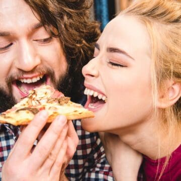 A man and a woman are sharing a slice of pizza. The man has long hair and a beard, and the woman has blonde hair pulled back. Both are smiling and laughing. The background is blurry, suggesting an indoor setting.