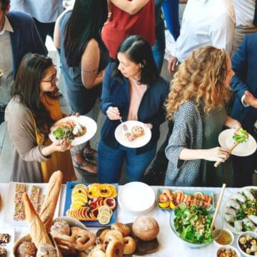 A group of people gather around a table filled with various dishes, including salads, bread, and fruit. Some guests hold plates with food, while others engage in conversation. The setting appears to be a social or corporate event.
