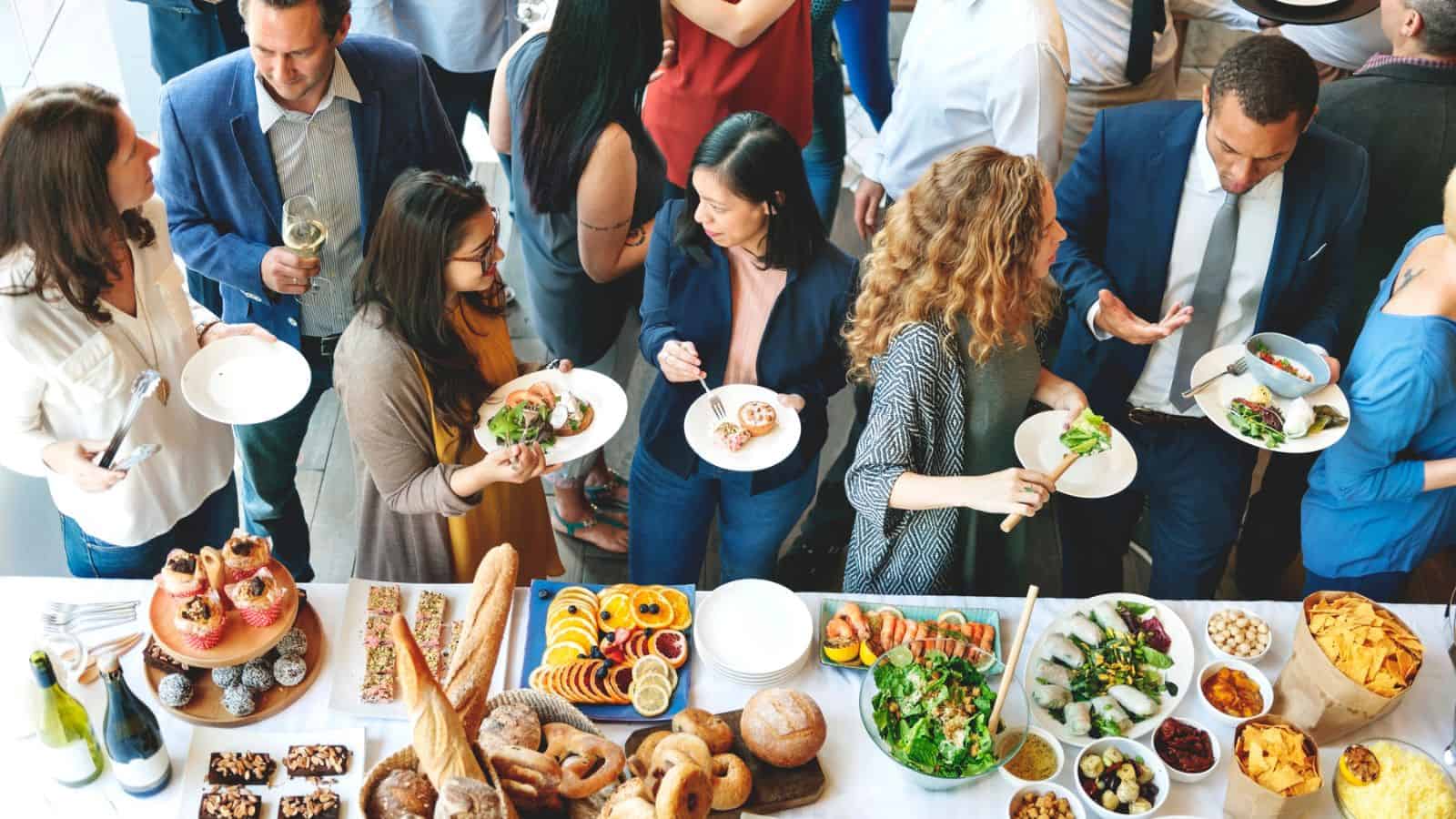 A group of people gather around a table filled with various dishes, including salads, bread, and fruit. Some guests hold plates with food, while others engage in conversation. The setting appears to be a social or corporate event.