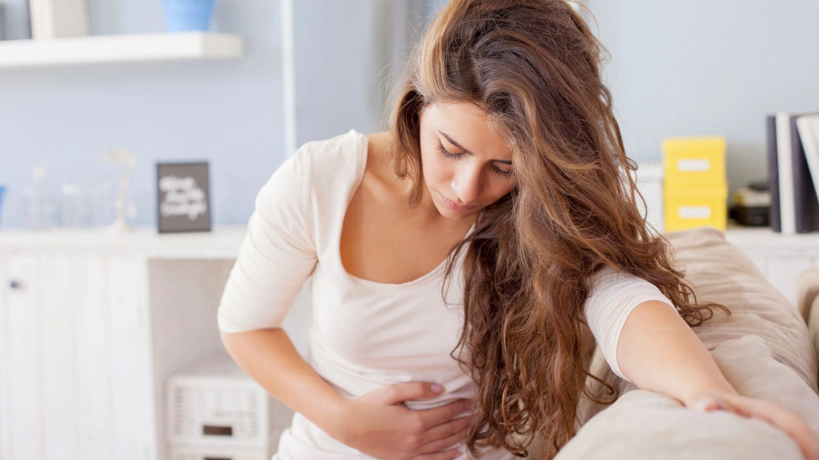 A woman with long brown hair wearing a white top leans on a couch, holding her abdomen with one hand. She appears to be in discomfort. The room is bright, with shelves and decorations in the background.