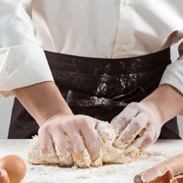 A person wearing a white shirt and black apron kneads dough on a floured surface. Eggshells, whole eggs, and a rolling pin are on the table.