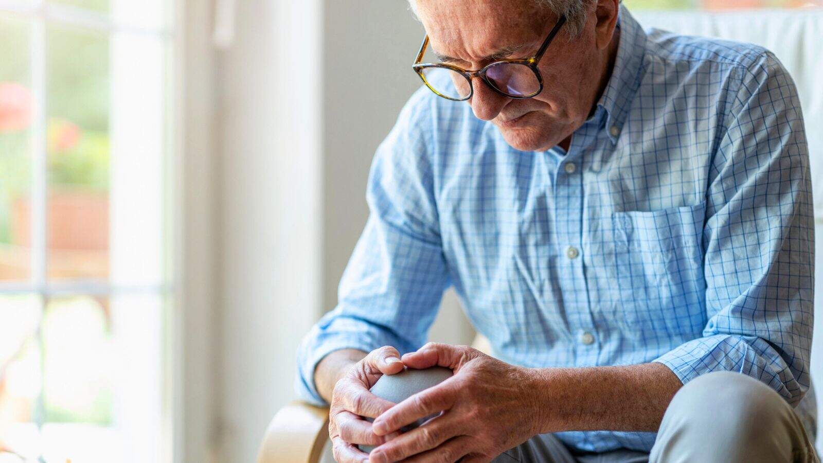 An elderly man in a light blue checkered shirt and glasses sits indoors, holding his knee with both hands. He appears thoughtful or concerned. A window in the background lets in natural light.