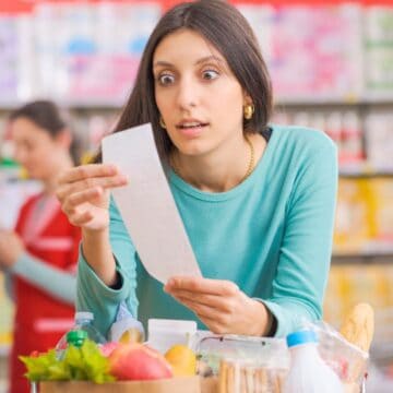 A woman with long dark hair looks surprised while examining a receipt in a grocery store. She is pushing a shopping cart filled with various groceries. Shelves stocked with products are visible in the background. Another person is visible further back.