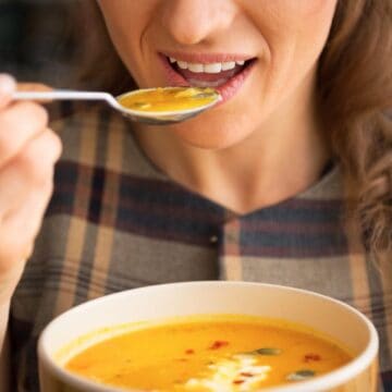 A person is holding a spoonful of orange soup close to their mouth, ready to take a bite. They are also holding a bowl filled with the same soup, which appears to be pumpkin or squash soup, garnished with seeds and a drizzle of cream.